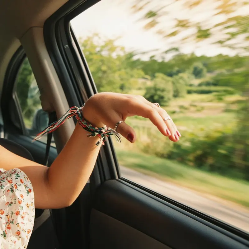 A hand with silver rings, painted nails and woven friendship bracelets trailing out of the open window of a moving car in bright sun, a bare sunlit arm and the edge of a summer dress in frame, green summer landscape rushing past.