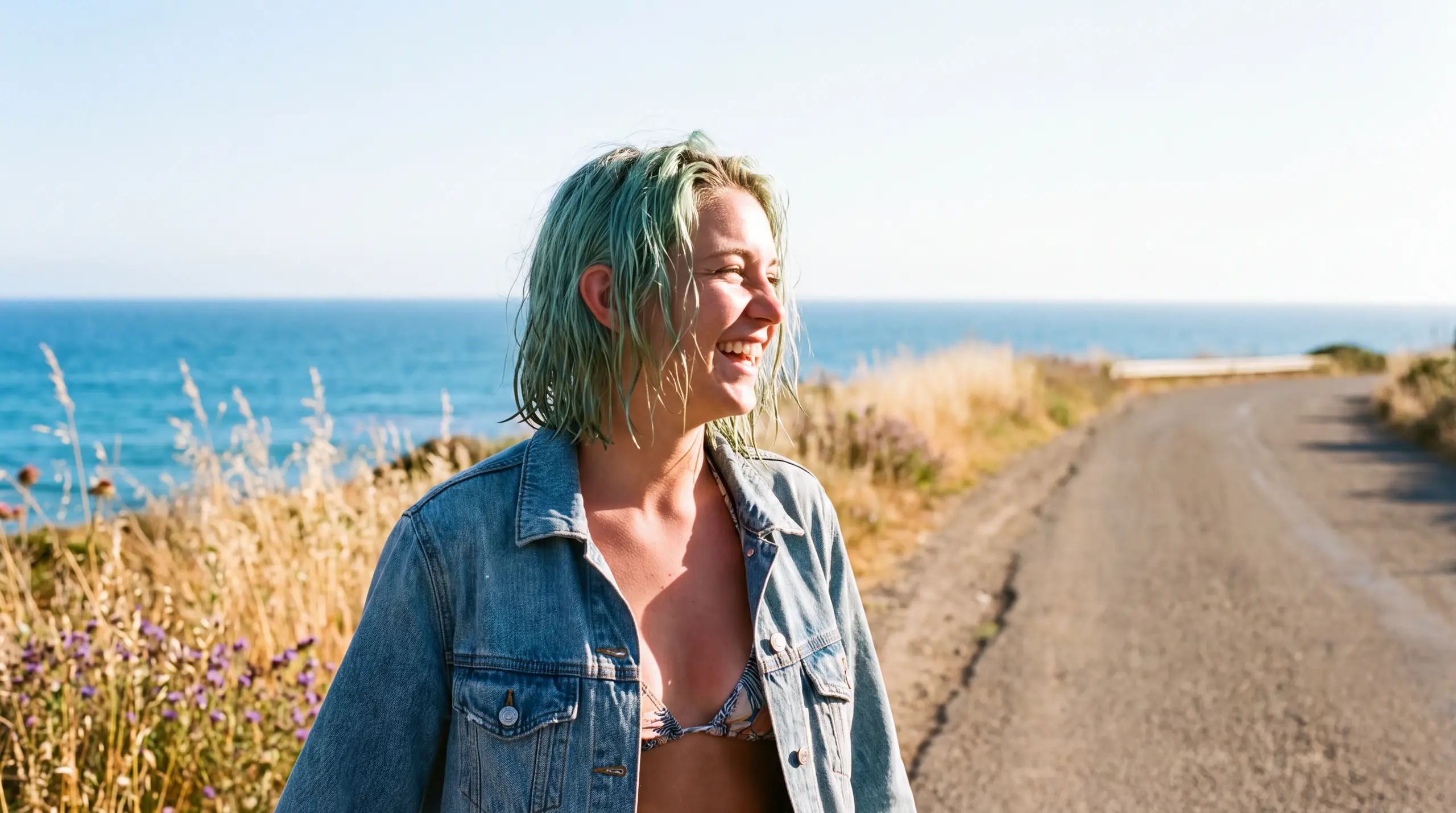 A woman in her early twenties with a wet seafoam green bob and a denim jacket worn open over a bikini top, walking a hot coastal road in full late-morning sun, sunburnt shoulders and salt drying on her skin, wildflowers and tall grass along the verge, bright open sky and blue water beyond, nothing in her ears, head turned away from the camera, laughing at something she is hearing.