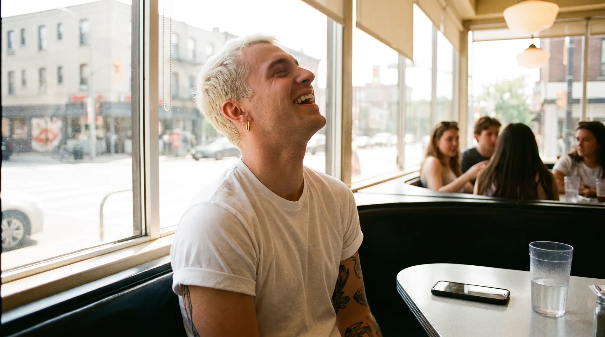 A man in his mid-twenties with platinum dyed hair, a gold earring and a tattooed forearm in a bright diner booth at midday, a white tee with the sleeves pushed up, big windows behind him, head back mid-laugh, nothing in his ears, phone face-down on the table.